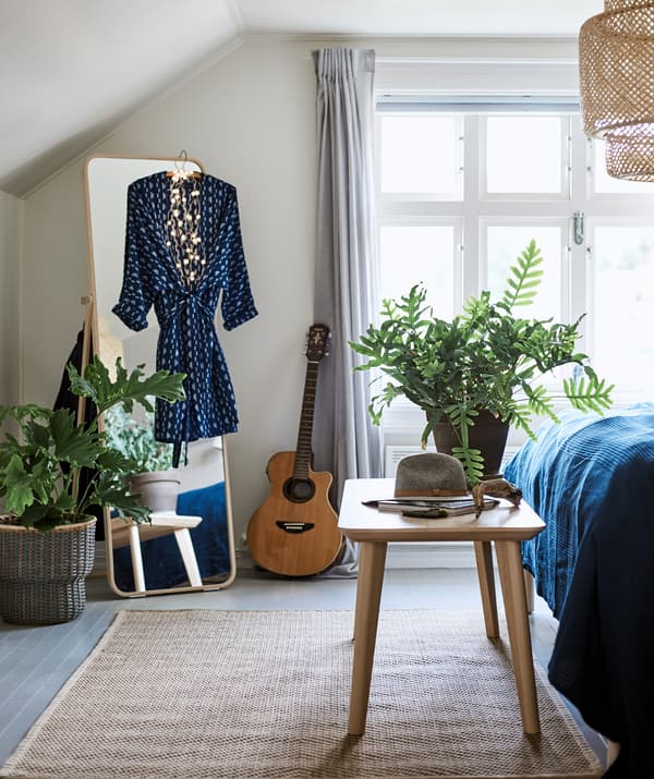 ห้องนอนที่มีโคมไฟติดเพดานพร้อมโป๊ะโคมสานและพรมสาน กระจกเงาตั้งอิสระมีชุดเดรสแขวนอยู่ และมีม้านั่งที่ปลายเตียง