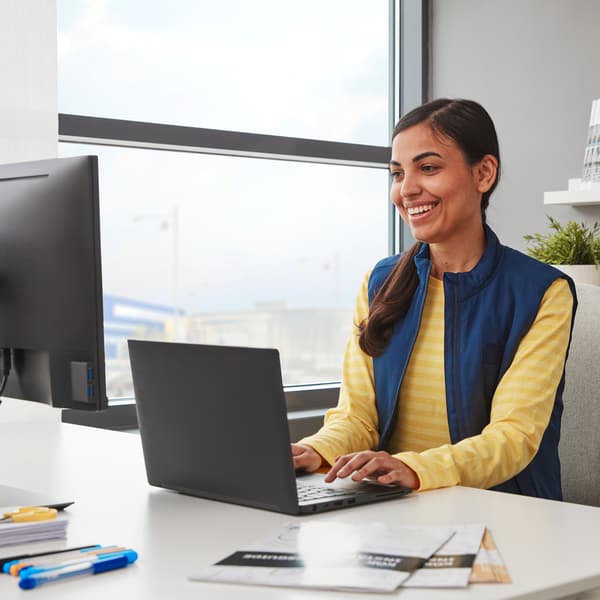 בחורה חייכנית בסוודר צהוב ואפוד כחול עובדת על מחשב נייד שחור המוצב על שולחן כתיבה לבן ליד חלון.