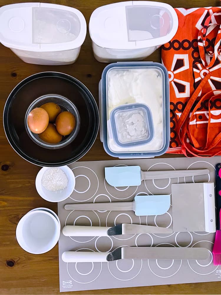 שולחן עץ במבט מלמעלה, שעליו שלל מכלים שונים עם מצרכים לאפייה כמו קמח, סוכר וביצים. עוד פריטים שיש על השולחן: כלי אפייה כמו מריות מגומי ומריות מדורגות.