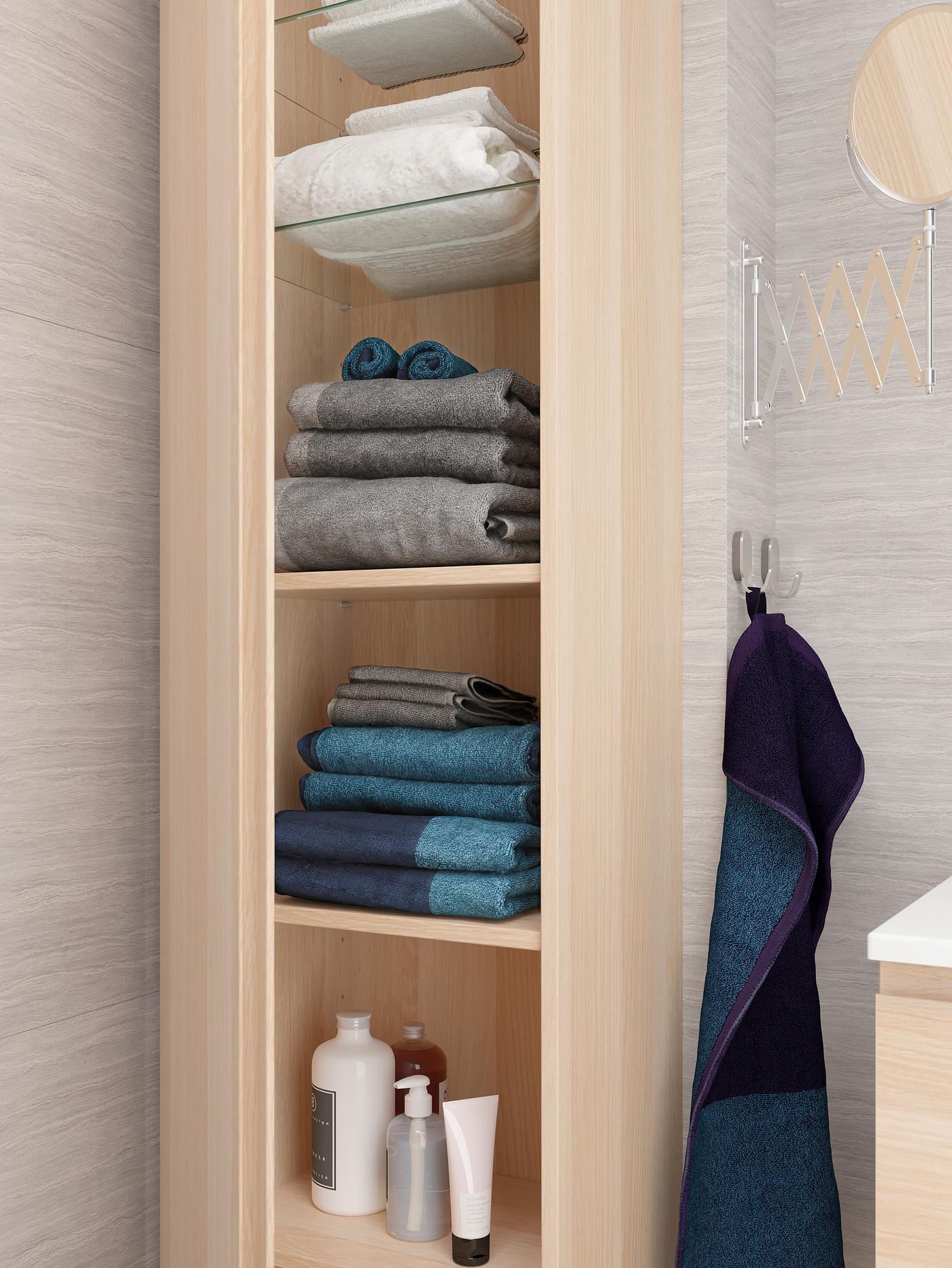 ארונית גבוהה עם אפקט אלון לבן, הארונית פתוחה ובתוכה מגבות ומוצרי רחצה על מדפי זכוכית ועץ.