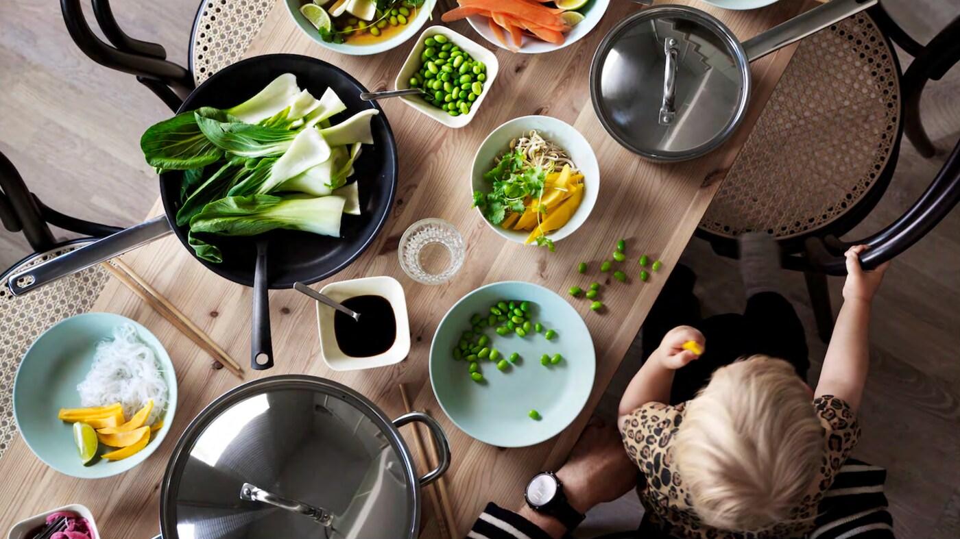 เด็กนั่งตักผู้ใหญ่ กำลังทานอาหารที่ใช้มือหยิบทานได้อยู่ที่โต๊ะไม้ซึ่งมีชามใส่ผักผลไม้และกระทะวางอยู่