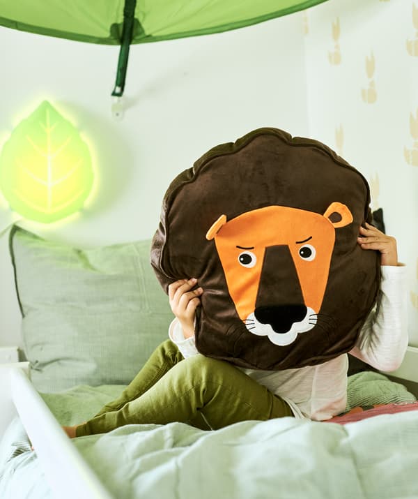 เด็กนั่งบนเตียงใต้มุ้งคลุมเตียงรูปใบไม้ ในมือถือหมอนอิงรูปสิงโตปิดหน้าตัวเองไว้