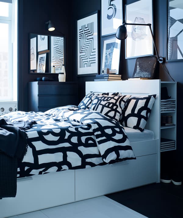 ห้องนอนสีดำและสีขาวโทนเดียวพร้อมเตียงที่มีที่เก็บของแบบบิวท์อิน และเครื่องนอนลายกราฟิกกับงานศิลปะบนผนัง