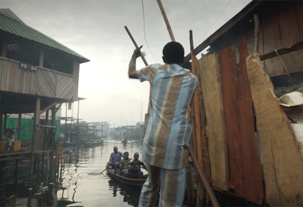 ผู้ชายในชุดหลายสีถ่อเรือผ่านเขตชุมชุนแออัดที่บ้านสร้างด้วยไม้ติดริมน้ำ