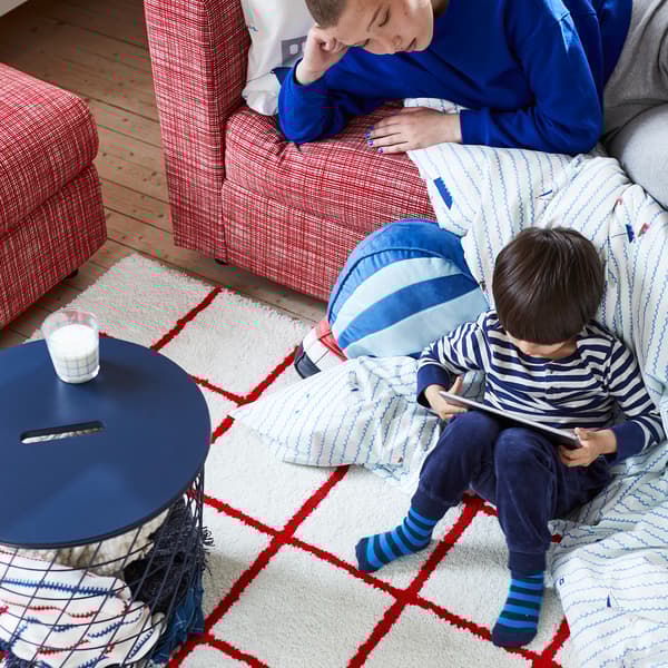 เด็กผู้ชายนั่งบนปลายผ้านวมที่พาดลงมาจากโซฟา ในมือถือแทบเล็ต มีผู้ใหญ่ที่อยู่บนโซฟามองอยู่