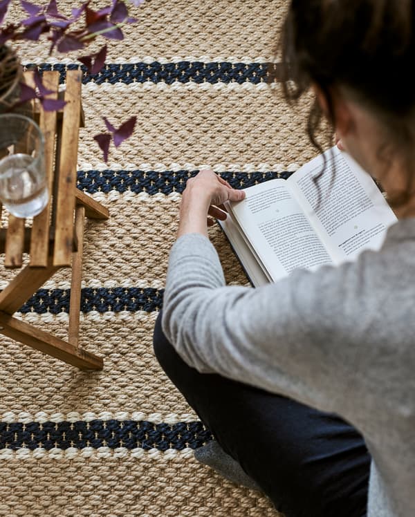 ผู้หญิงนั่งอ่านหนังสือบนพรมถักลายทางทำจากเส้นใยธรรมชาติ มีโต๊ะข้างพับได้ ต้นไม้ และแก้วน้ำ