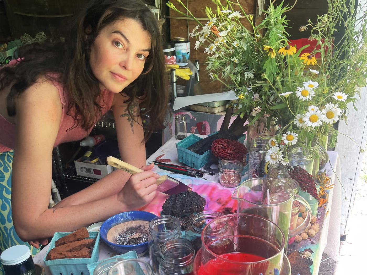 אישה נשענת על שולחן כתיבה מלא בצנצנות זכוכית ופרחים. היא משתמשת במכחול כדי לצבוע פיסת בד בצבע ורוד.