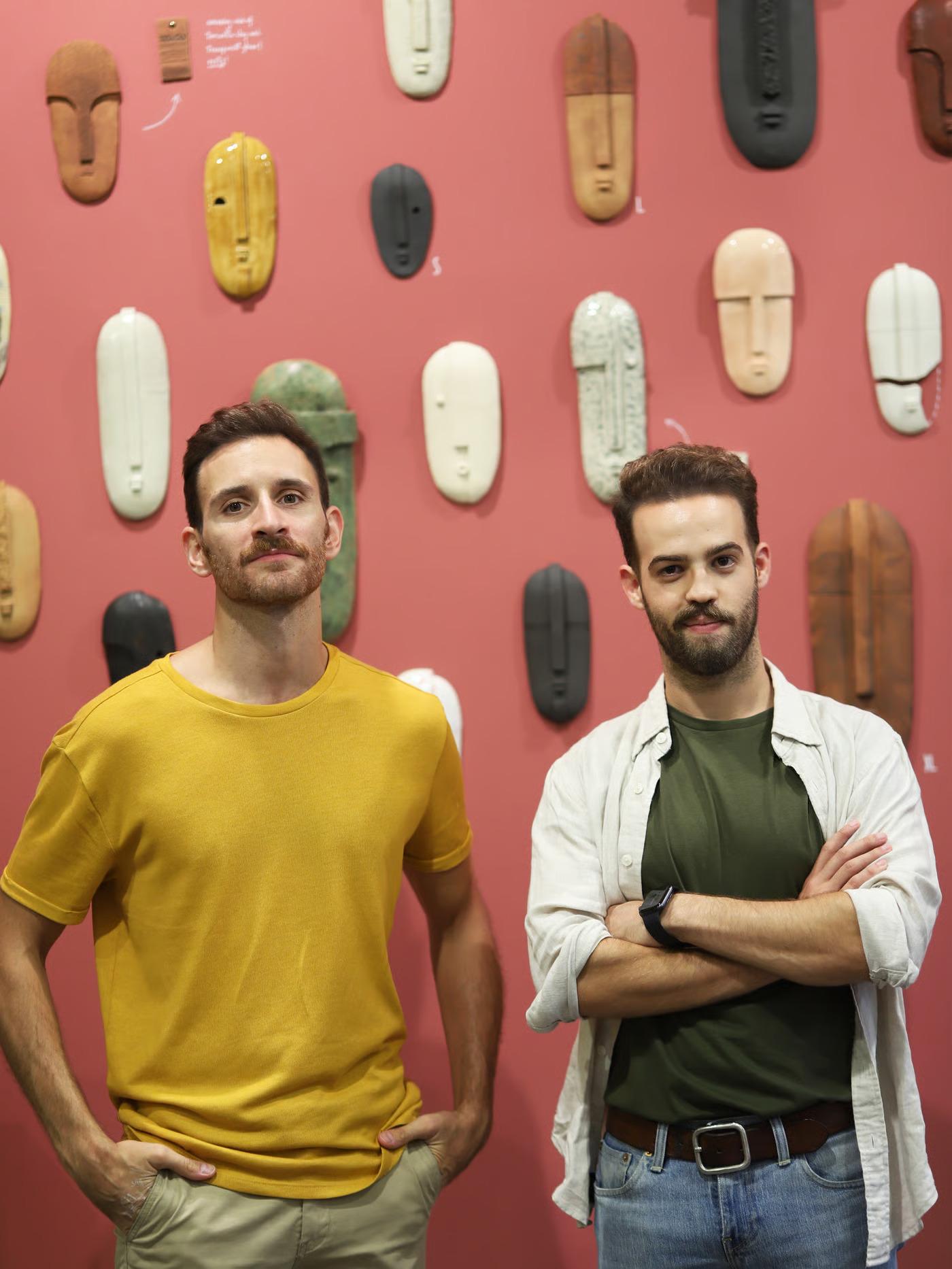 שני גברים צעירים עומדים מול קיר ורוד שמעוטר במסכות חמר בצבעים שונים.