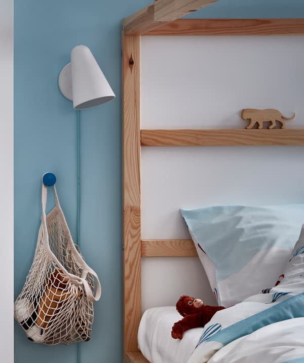 ด้านหัวเตียงของเตียงเด็กมีถุงตาข่ายใส่ตุ๊กตาผ้าแขวนบนมือจับประตูข้างเตียง โคมไฟติดผนังและตุ๊กตาผ้าข้างหมอน