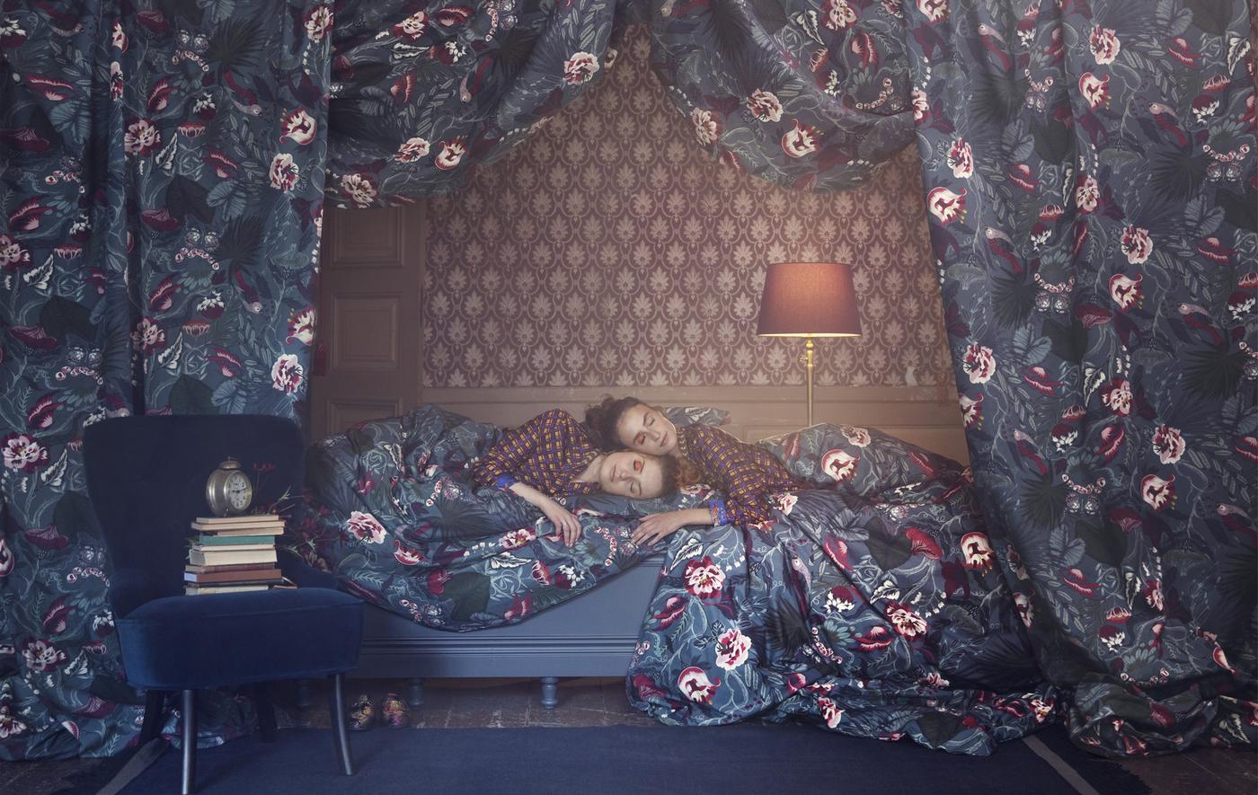 หญิงสองคนนอนบนเตียงโดยมีวอลเปเปอร์ที่มีลวดลายล้อมรอบด้วยผ้าลายดอกไม้