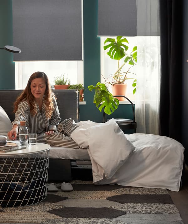 ผู้หญิงสวมชุดนอนนั่งบนโซฟาเบดที่ใช้เป็นเตียงนอน แก้วน้ำอยู่บนโต๊ะกลาง ม่านม้วนทึบแสงที่ปิดไว้ครึ่งหนึ่งอยู่ด้านหลัง