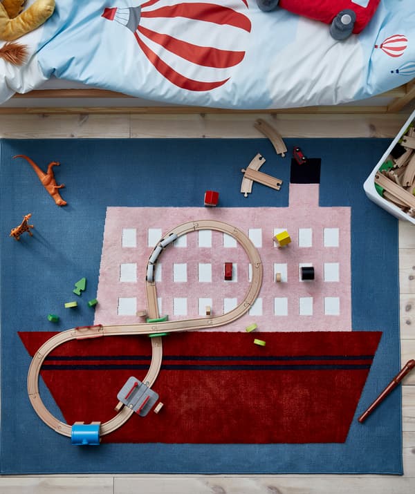 พรมลายเรือปูอยู่ตรงพื้นข้างเตียงนอนของเด็ก ชิ้นส่วนรางรถไฟที่ทำด้วยไม้และของเล่นชิ้นอื่น ๆ วางกระจายอยู่