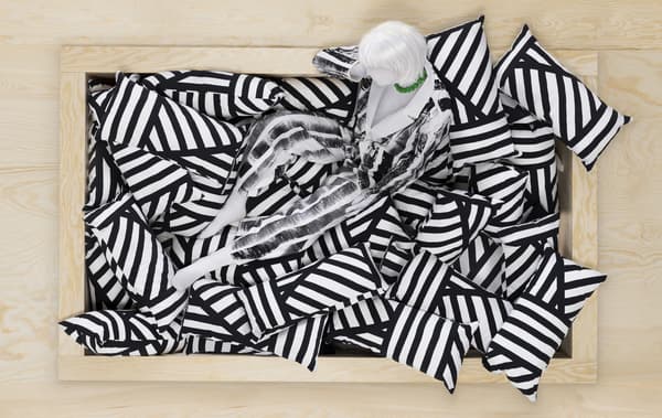 หุ่นจำลองนั่งอยู่บนกองหมอนอิงลายแถบสีขาวและสีดำในกรอบไม้