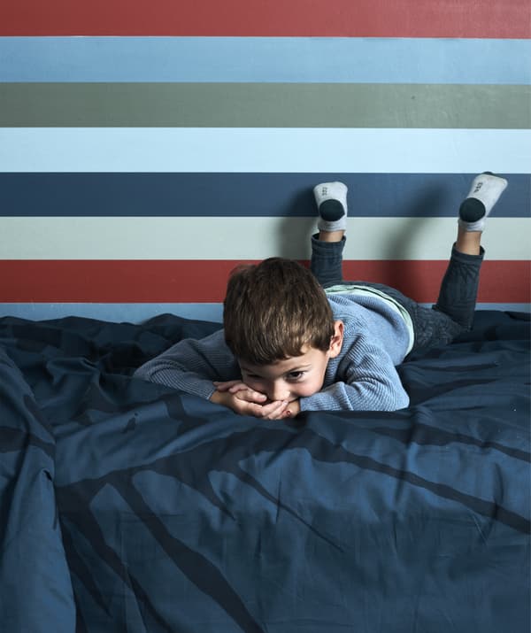 เด็กชายนอนบนเตียงที่มีเครื่องนอนสีน้ำเงินเข้มและวอลเปเปอร์ลายทางอยู่ด้านหลัง