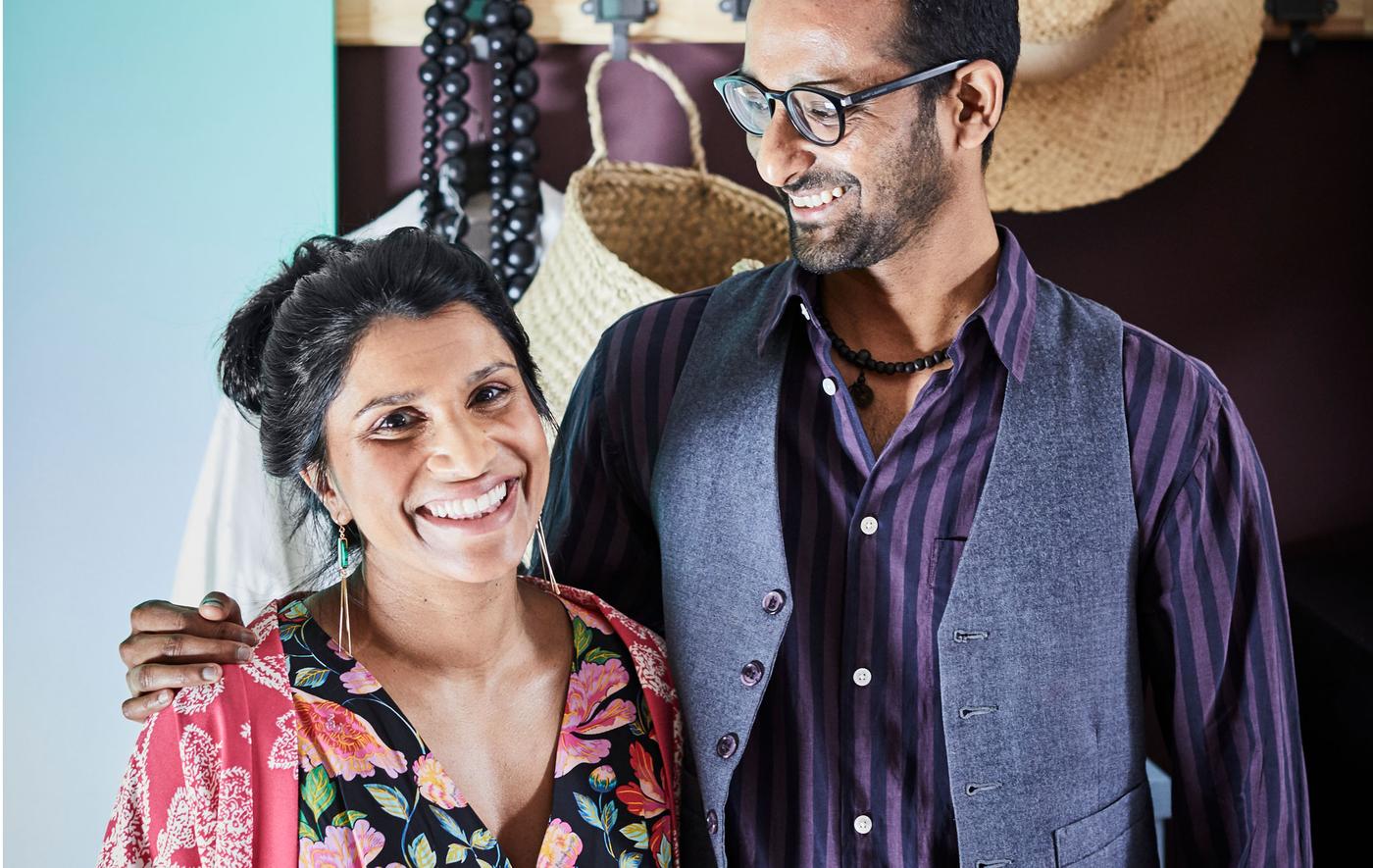 ผู้หญิงและผู้ชายอายุประมาณสามสิบยืนยิ้มอยู่ด้วยกันด้วยความรัก มีเสื้อผ้าและของใช้อยู่ที่ชั้นด้านหลัง