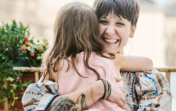 ผู้หญิงนั่งอยู่บนระเบียง สวมชุดกระโปรงลายตารางสีฟ้ายิ้มกว้าง กอดเด็กผู้หญิงสวมชุดสีชมพู