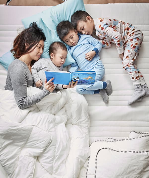 แม่และลูกสามคนนอนอ่านหนังสือด้วยกันบนเตียงที่มีเครื่องนอนสีขาว