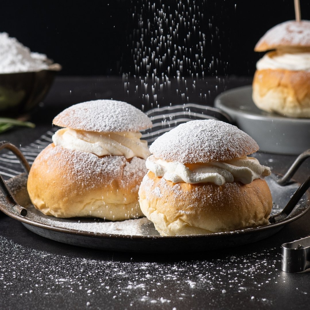 슈가 파우더가 눈처럼 흩날리는 가운데, 부드러운 크림을 가득 채운 스웨덴 전통 디저트 셈라가 검은 테이블 위에 놓여있는 이미지