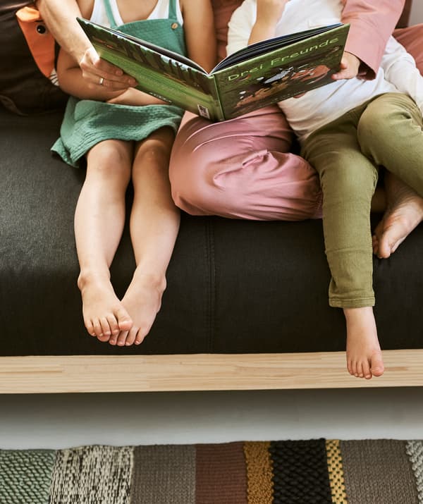 ผู้หญิงสวมชุดจั๊มสูทสีชมพูนั่งบนโซฟาสีเทาโครงทำด้วยไม้ กำลังอ่านหนังสือสำหรับเด็ก แขนโอบรอบเด็กผู้ชายและเด็กผู้หญิง