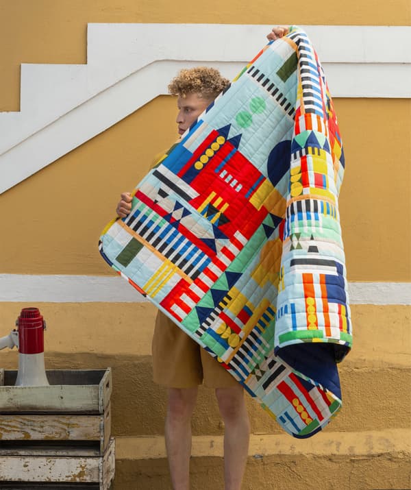 ผู้ชายถือผ้าห่มที่มีลวดลายหลากสีอยู่ด้านหน้ากำแพงสีเหลือง