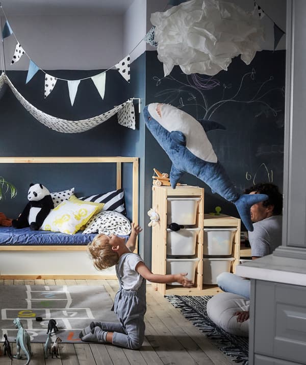 เด็กกำลังโยนตุ๊กตาผ้าปลาฉลามในอากาศ มีกล่องเก็บของ และมุ้งผ้าอยู่ด้านหลัง