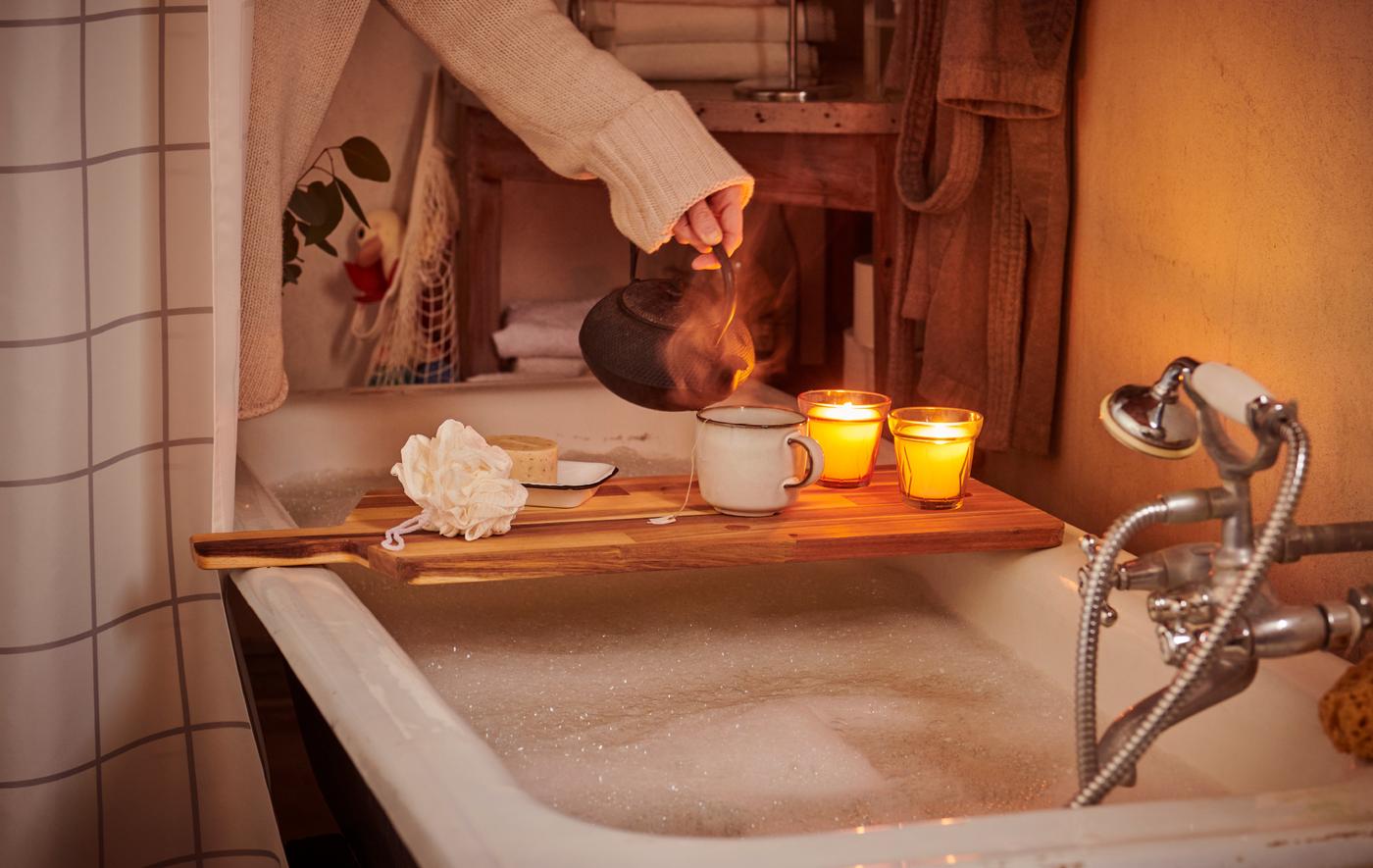 ผู้หญิงสวมเสื้อกันหนาวกำลังเตรียมทำสปาโดยเทน้ำชาลงในแก้วมัคที่อยู่บนเขียงไม้ซึ่ีงพาดอยู่บนอ่างอาบน้ำ