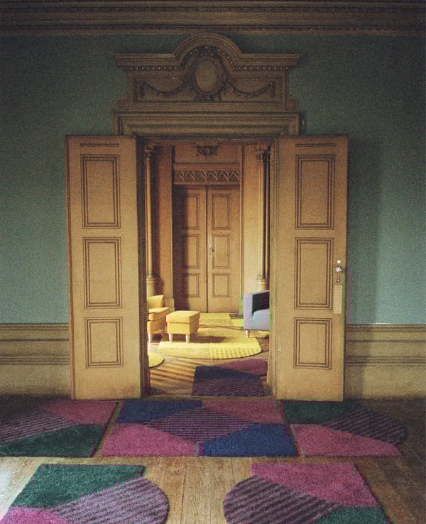 ประตูเข้าบ้านบานใหญ่พร้อมพรมที่มีลวดลายอยู่ด้านหน้า เปิดสู่ห้องนั่งเล่นที่มีเก้าอี้สีเหลือง สตูลวางเท้าและพรม