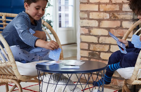 เด็กสองคนจดจ่อกับการเล่นการ์ดเกมบนอาร์มแชร์หวายคนละด้านโดยมีโต๊ะกลางคั่นอยู่