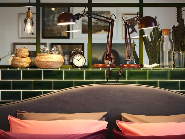 두 개의 다크레드 독서등이 그린 톤 타일의 반벽 위에 놓여 그레이 색상의 침대헤드와 해초 바구니, 베개 여섯 개를 비춰주는 모습.