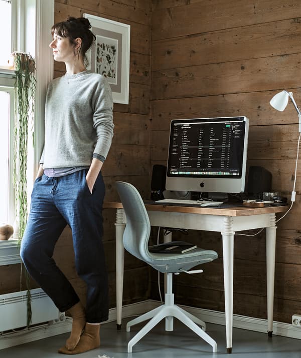 ผู้หญิงยืนมองออกไปนอกหน้าต่างตรงมุมห้องที่ทำเป็นโฮมออฟฟิศพร้อมโต๊ะทำงานและเก้าอี้หมุน