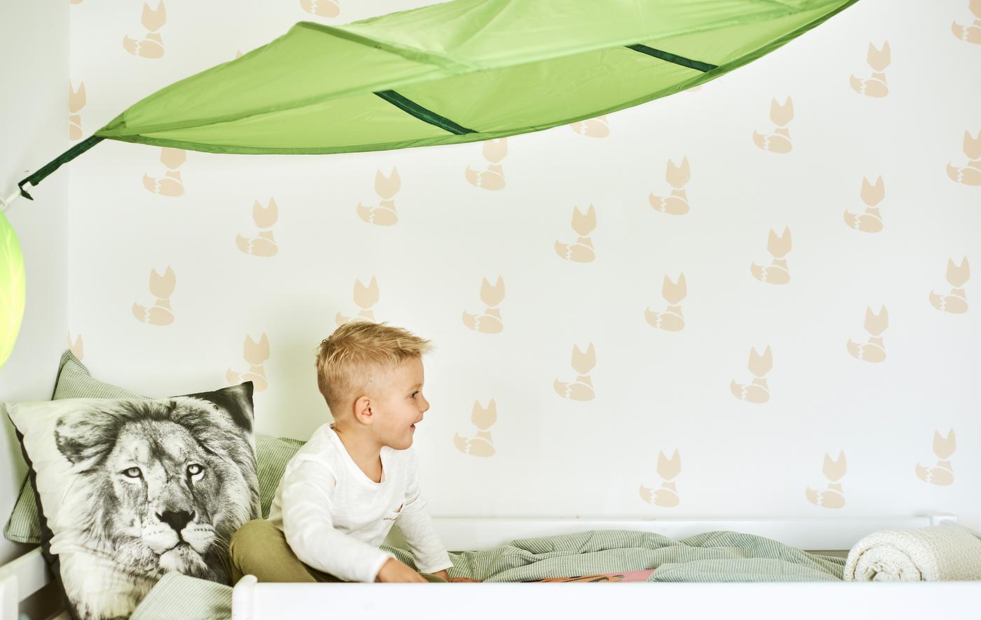 เด็กผู้ชายนั่งอยู่บนเตียงที่มีผ้านวมสีเขียวและหมอนอิงพิมพ์ลายหน้าสิงโต มีมุ้งคลุมเตียงรูปใบไม้ขึงไว้ด้านบน