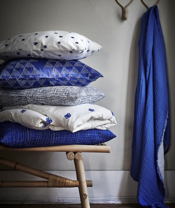 หมอนอิงลายน้ำเงินขาววางซ้อนกันอยู่บนม้านั่งหวาย และผ้าสีน้ำเงินแขวนอยู่บนตะขอ