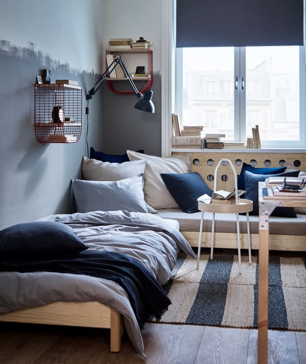 ห้องที่มีโซฟาเข้ามุมและเตียงเดี่ยว พร้อมสิ่งทอสีน้ำเงิน สีเทา และสีธรรมชาติ รวมทั้งและตะกร้าลวดสีแดงเก็บของติดผนัง