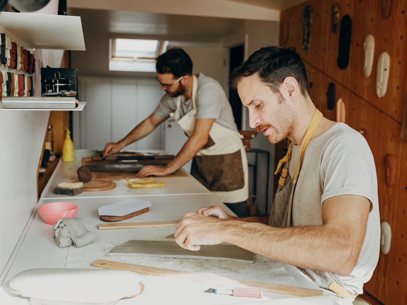 שני גברים צעירים בחולצות טריקו וסינרים יושבים ליד שולחן ומכינים כלים ויצירות אומנות מחרס.