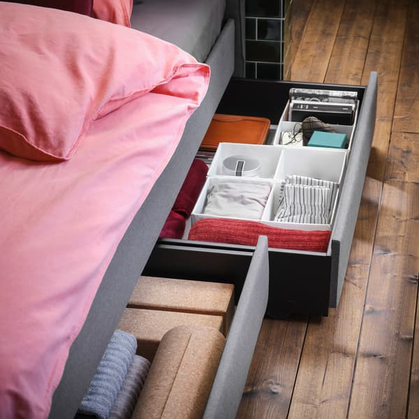 그레이 색상의 쿠션형 침대와 라이트브라운레드 색상의 침구, 오픈 수납서랍 두 개. 작은 소지품들이 정리되어 있는 수납함.