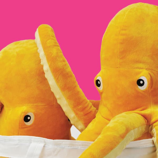 2 gule blæksprutte tøjdyr fra serien BLÅVINGAD stikker hovederne op af en hvid pose på en pink baggrund 