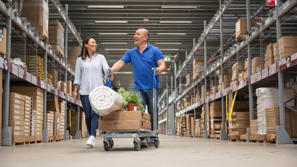 2 IKEA customers pushing a flat cart with boxes and a mattress through a warehouse.