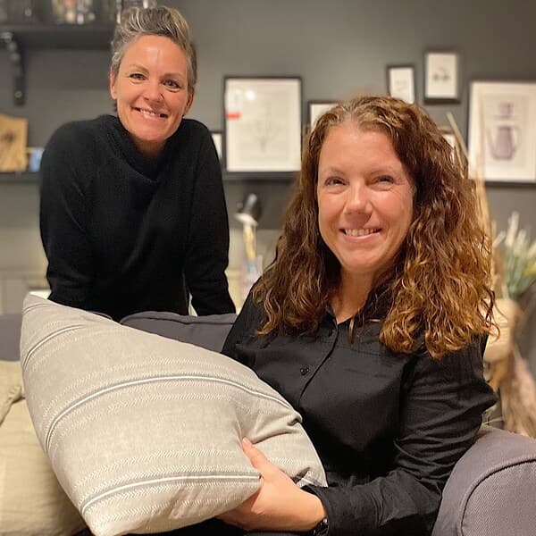 2 kvinder i en stue, hvor den ene sidder i en grå sofa med en pude i hænderne og den anden står i baggrunden.