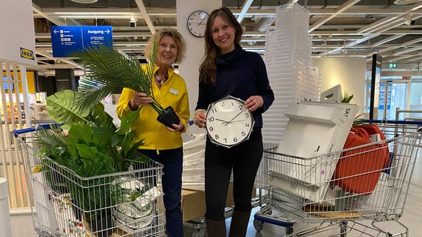 2 Personen lachen in die Kamera und halten verschiedene Einrichtungsgegenstände hoch. Eine IKEA Mitarbeiterin in gelber Jacke hält eine Grünpflanze in der Hand. Die andere Person hält eine Wanduhr. Im Hintergrund sind IKEA Einkaufswägen mit weiteren Dekoartikeln zu sehen.
