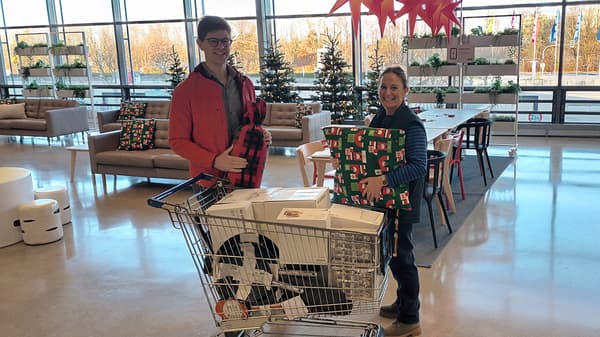 2 Personen stehen im weihnachtlich dekorierten IKEA Einrichtungshaus hinter einem Einkaufwagen randvoll mit Geschirr, Besteck, Töpfen und weiteren Kochuntensilien. Eine davon trägt ein blaues IKEA Mitarbeitenden T-Shirt.  