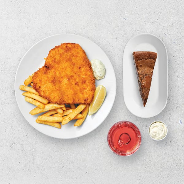 2 white plates, a drink, and a sauce pot on a grey concrete background with IKEA chicken schnitzel and chips, with a slice of cake and a glass of juice