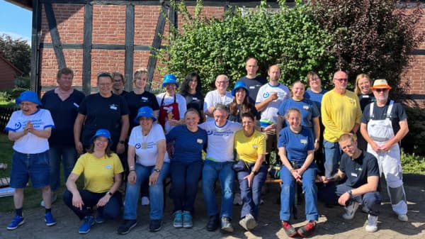 22 fröhlich lachende Personen, teils in Arbeitskleidung, stehen vor einem alten restauriertem Backstein-Fachwerkhaus. 
