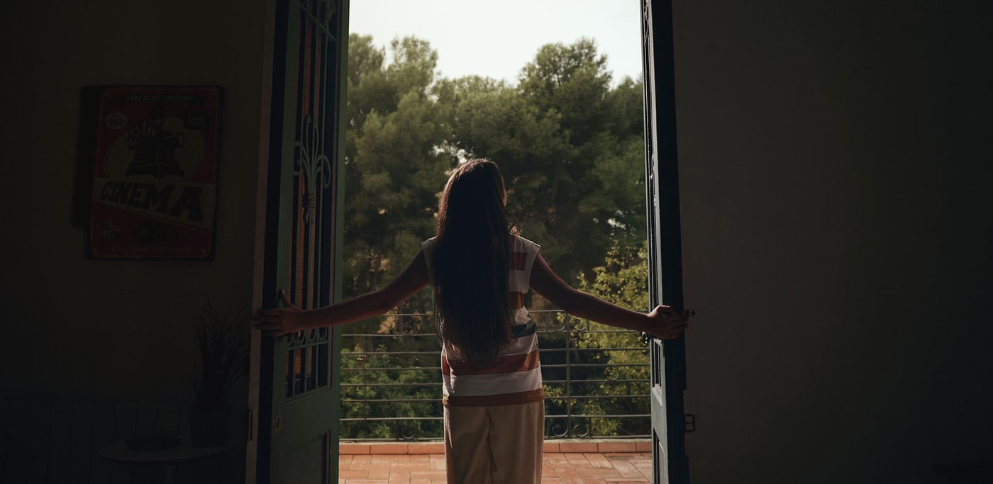 A woman with a red and white striped shirt holding doors open leading to an ourside space with trees in the background.
