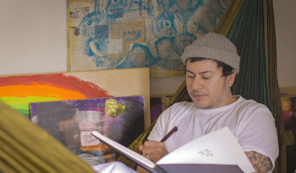 2SLGBTQ+ campaigner, Fox Fisher, is sitting in a hammock, surrounded by pieces of art. He is writing in a sketchbook.