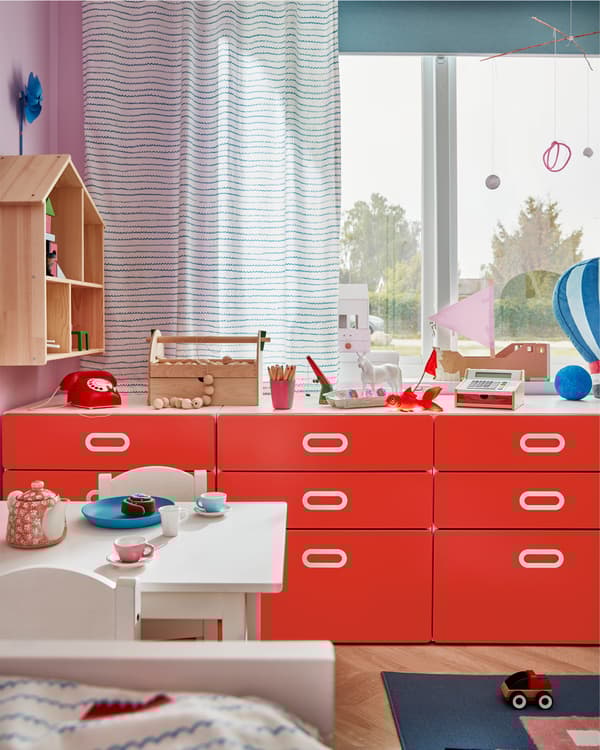 창문 앞에 레드 서랍장 3개가 놓여 있는 아이 방, 서랍장 상판에는 장난감과 놀이용품을 놓을 수 있는 공간이 마련되어 있는 모습