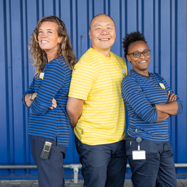 3 IKEA Mitarbeiter:innen, die Schulter an Schulter vor einer blauen Wand stehen