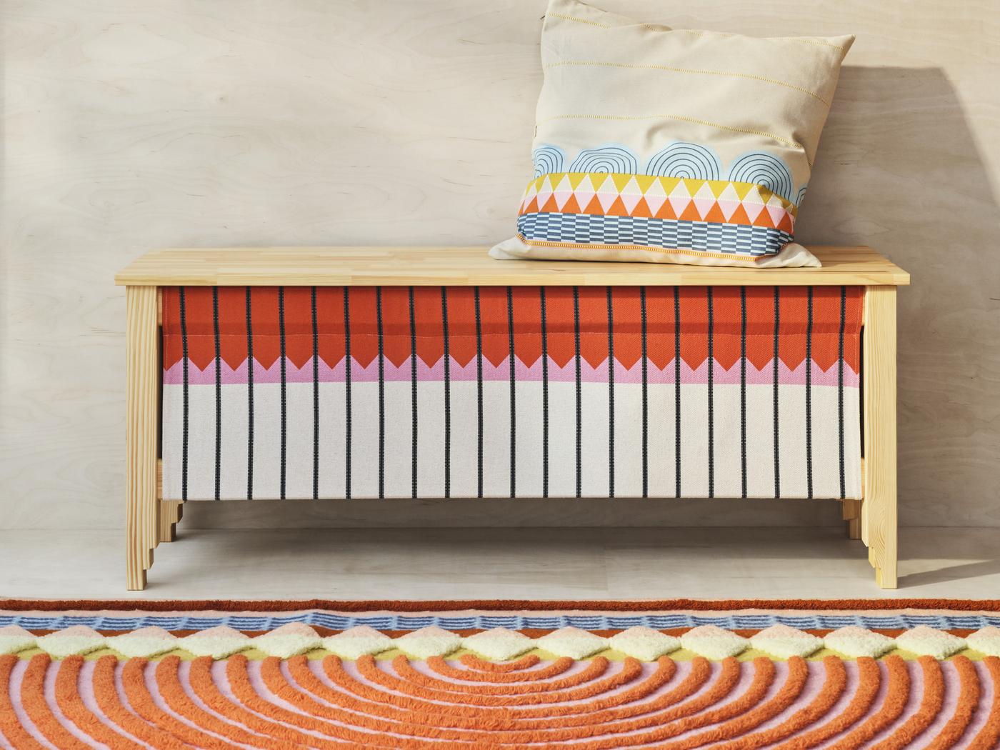 3 Produkte der neuen OMMJÄNGE Kollektion, ein farbenfroher Teppich in Form eines Regenbogens, eine Kiefernholz Bank, bunt bemalt und ein Kissenbezug mit buntem Muster in hellblau und orange