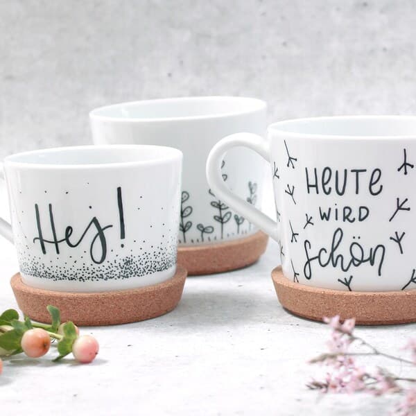 3 weiße Tassen mit einem grünen Muster und einer grünen Schrift stehen auf einem weißen Tisch.