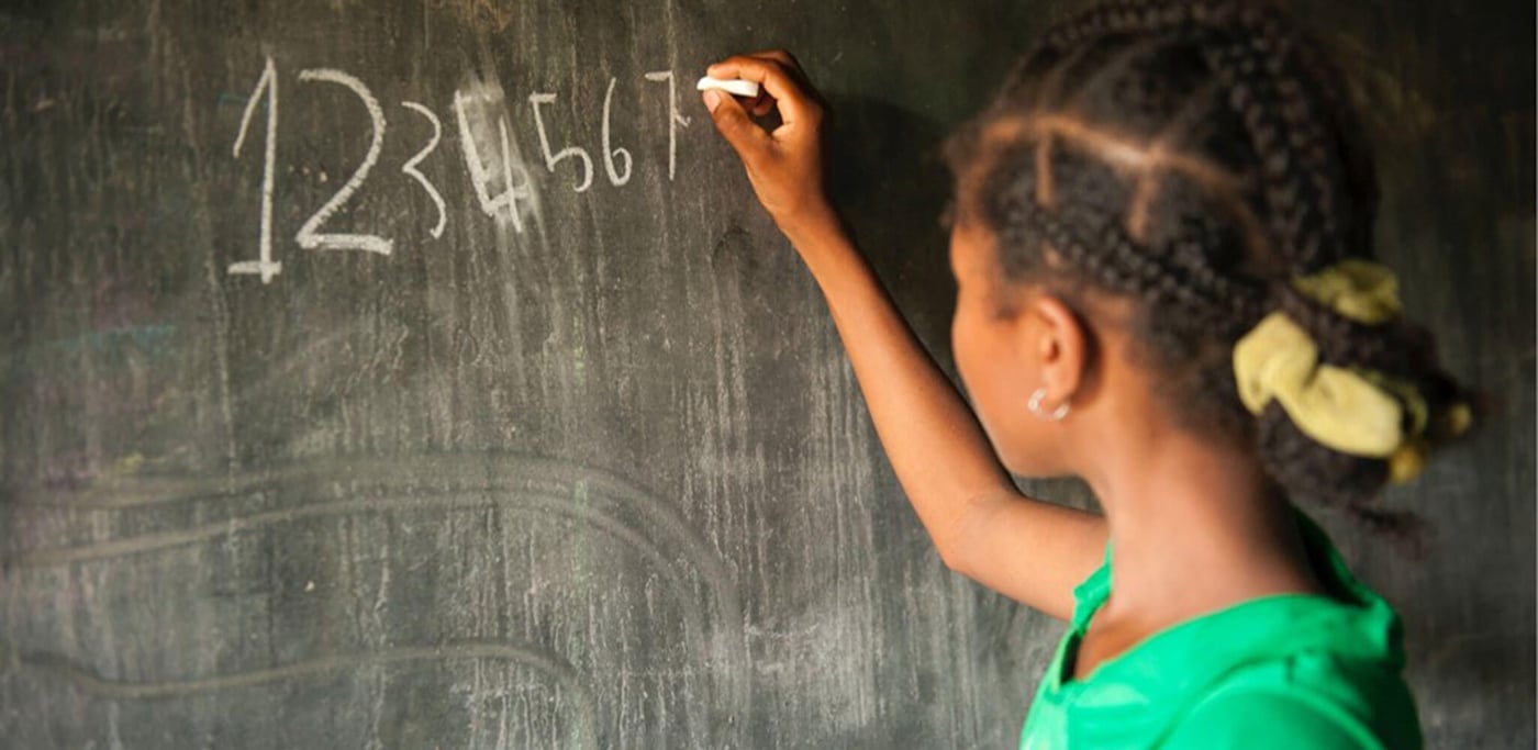 A young girl facing a chalkboard writing her numbers on the board from 1 to 7