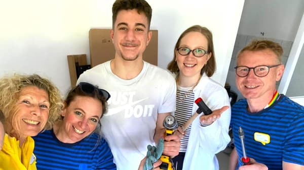 5 Personen lachen in die Kamera und halten verschiedene Werkzeuge in der Hand. In der Mitte steht ein junger Mann und sieht sehr glücklich aus.
